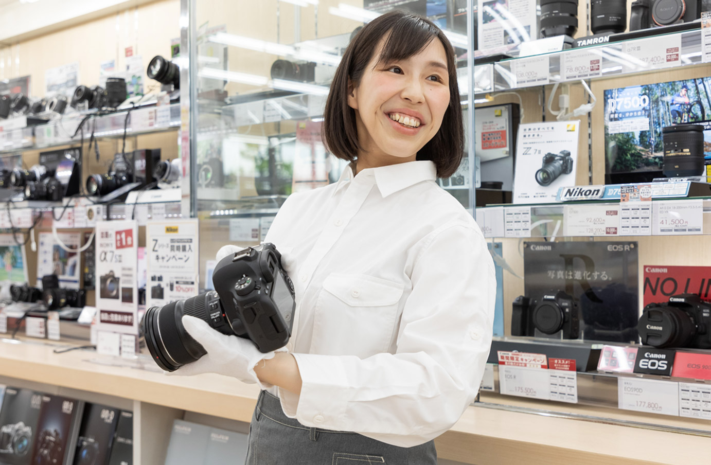 カメラが陳列された商品棚の前で一眼レフカメラを手に持ち明るく接客するカメラ販売店女性スタッフ